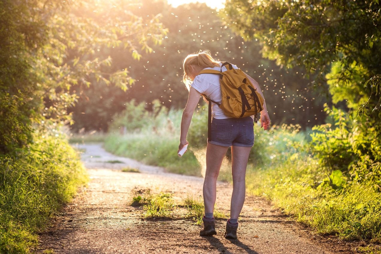 Woman on dirt road surrounded in trees -- and mosquitos spraying herself with insect repellent.